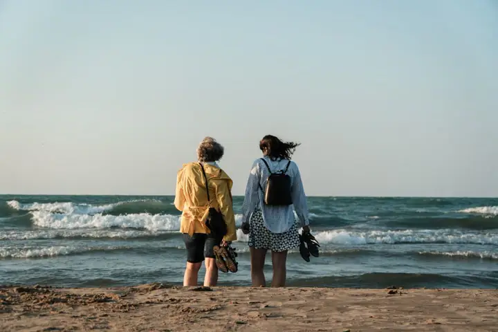 Duas pessoas de costas, uma de capa amarela e outra de mochila, lado a lado na areia olhando o mar.