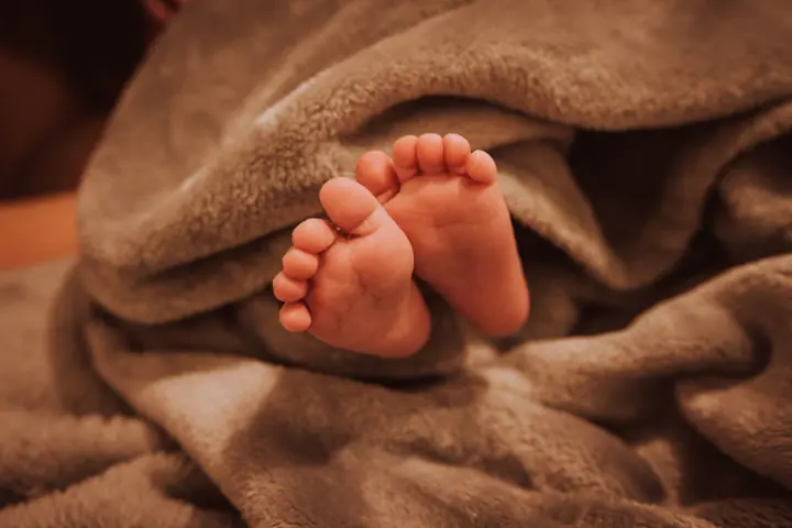 Dois pés de bebê aparecendo no meio de uma manta marrom, sem mostrar o rosto.