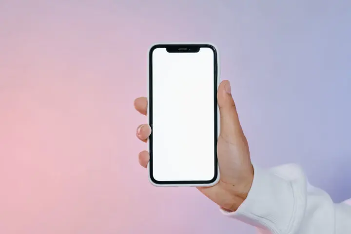 Uma mão adulta segurando um celular com a tela em branco, diante de um fundo liso.